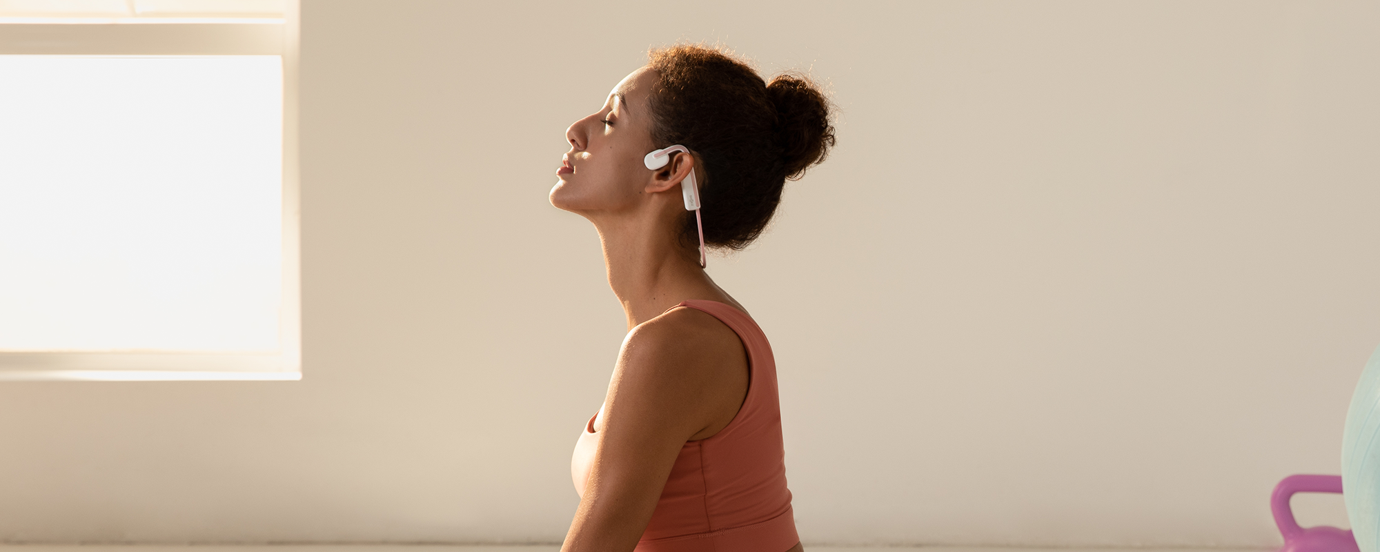 Image of a woman in an empty, well-lit room with eyes closed and wearing Shokz OpenMove headphones