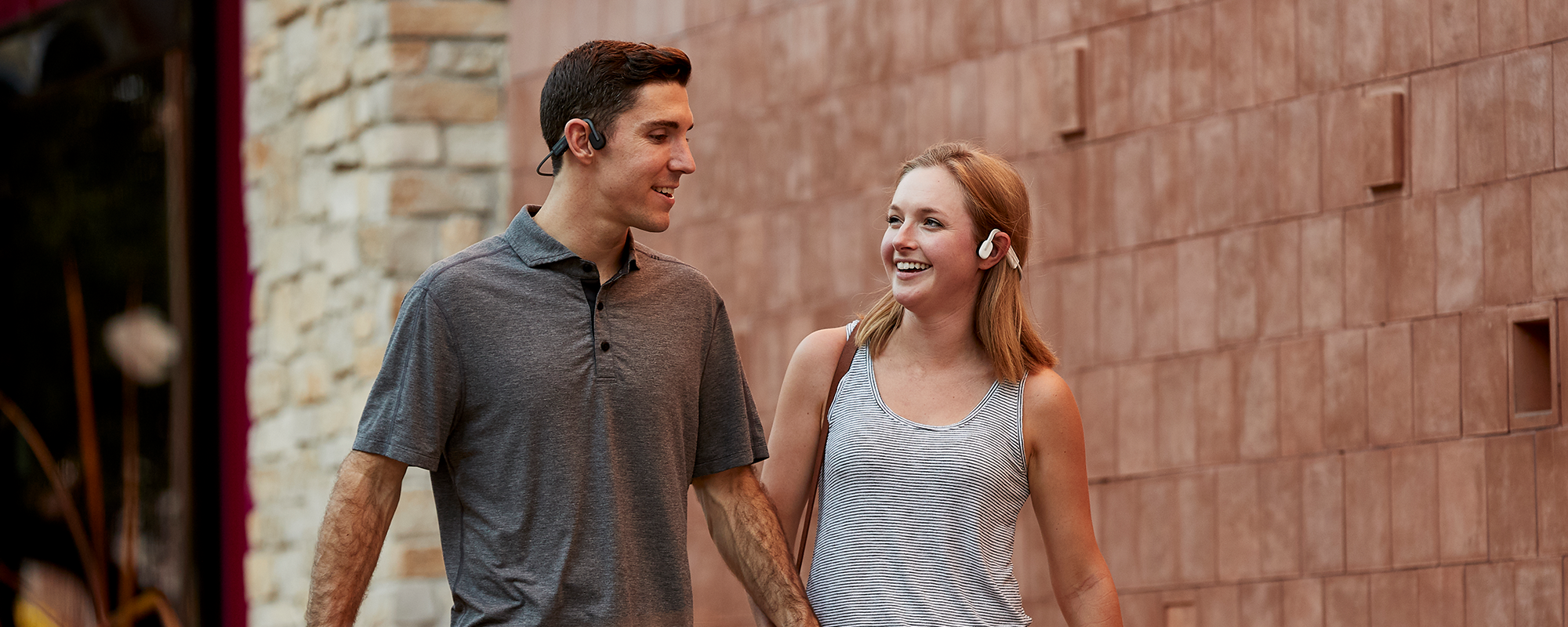 Man and woman walking on city sidewalk and both wearing Shokz OpenMove wireless headphones